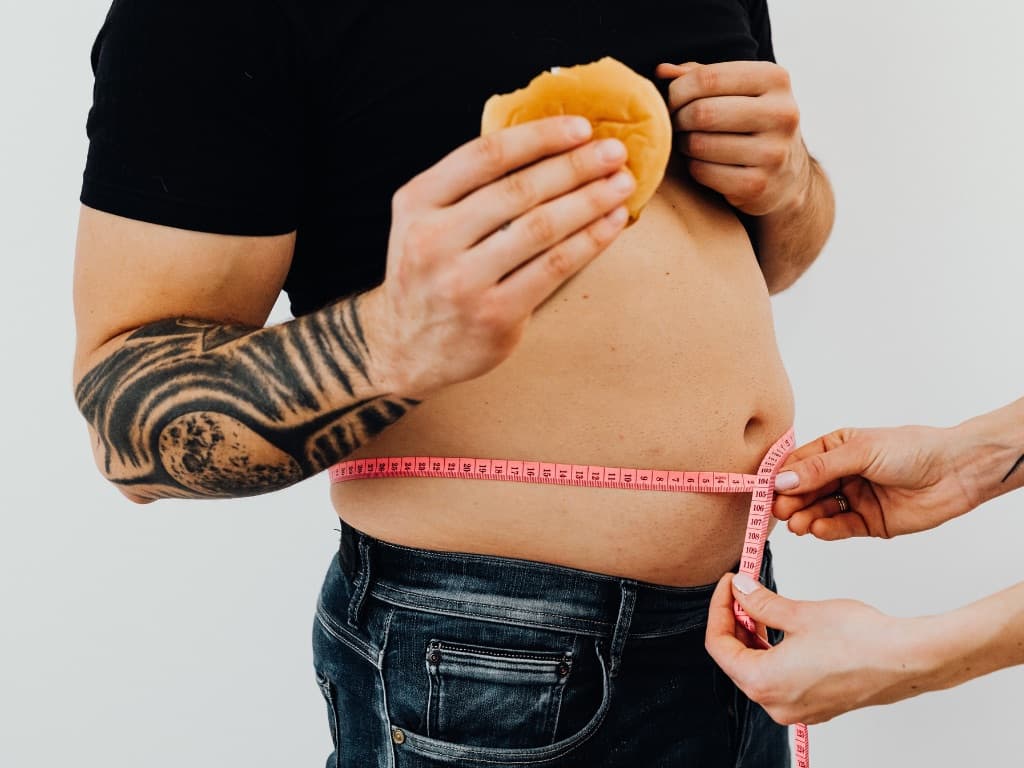 Man eating a cheeseburger while his waistline is being measured