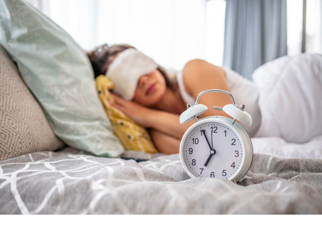woman sleeping in bed with a sleep mask and an alarm clock next to her on the bed